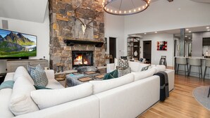 A modern living room with a stone fireplace, a large sofa, a flat-screen TV, and a dining area with bar stools.