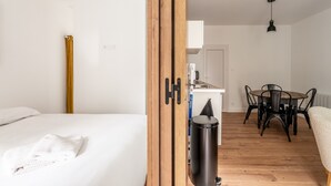 A room with a bed, a dining table with chairs, a trash bin, and a door with a wooden frame. - Holiday Home 'L'écrin De La Sèvre' with Wi-Fi (Cugand)