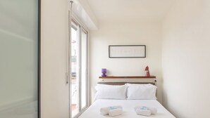 Habitación de hotel con una cama, una ventana, un cuadro en la pared y un pequeño estante con una lámpara morada y un objeto rojo.