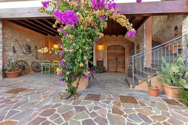 A stone-floored courtyard with a wooden staircase, potted plants, and a rustic stone wall.
