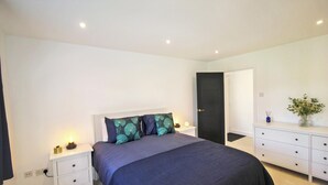 Un dormitorio moderno con una cama grande, mesitas de noche blancas, un aparador y una puerta que da a otra habitación.
