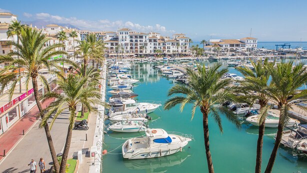 Ein Yachthafen mit zahlreichen vertäuten Booten, umgeben von Palmen und Gebäuden im mediterranen Stil.