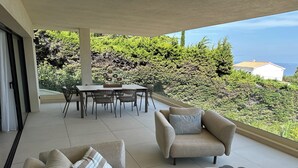 A modern outdoor living area with a dining table and chairs, a sofa with a striped pillow, and a view of a lush green hillside. - Villa Les Issambres, infinity pool, sea view (Roquebrune-sur-Argens)