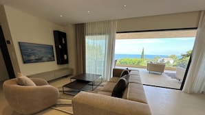 A modern living room with a sofa, a coffee table, and a large window offering a view of the ocean. - Villa Les Issambres, infinity pool, sea view (Roquebrune-sur-Argens)