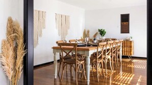Un coin repas avec une table et des chaises en bois, un mur blanc orné de décorations en macramé et une fenêtre avec des stores.