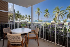 Ein Balkon mit rundem Tisch und Stühlen, der Blick auf einen Strand mit Palmen und einem klaren Himmel freigibt.