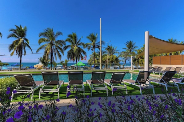 Un espace aménagé au bord de la piscine, avec des chaises longues, une piscine, des palmiers et une vue sur l’océan.