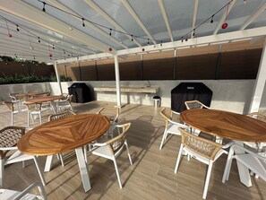 Eine Außenpatio mit runden Holztischen und Korbstühlen, einem Grill und einem überdachten Bereich mit Lichterketten.