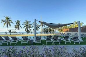 Ein Poolbereich mit Liegestühlen, einer beschatteten Markise und Blick auf das Meer.