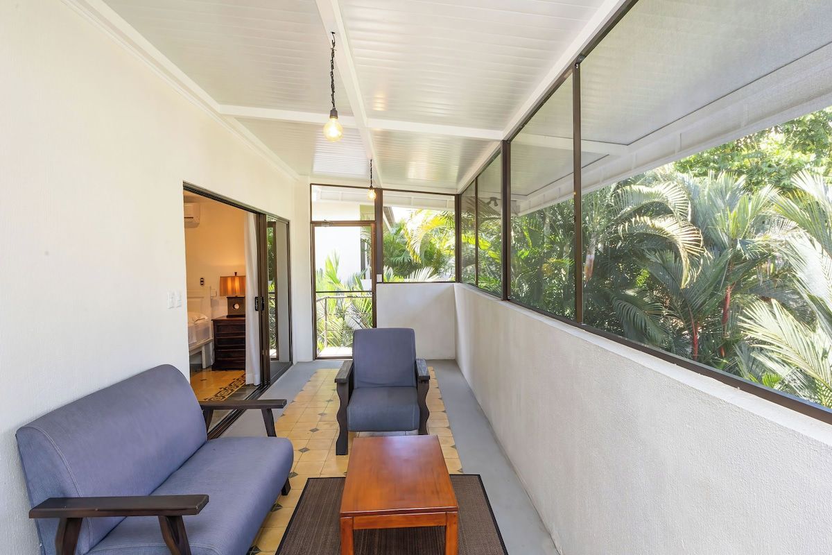 A balcony with a sofa, chairs, and a table, overlooking a lush garden.
