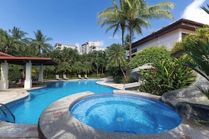 A swimming pool with a hot tub, surrounded by palm trees and lounge chairs, with buildings in the background. - Jaco Beach House (Jaco)