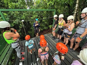 Eine Gruppe von Menschen auf einer Holzplattform mit Sicherungsgeschirren, Helmen und Seilen, was auf eine Ziplining-Aktivität hindeutet.