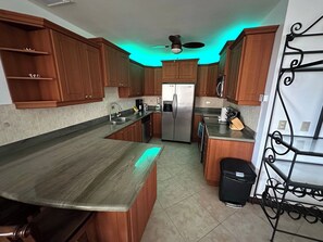Eine Küche mit Holzschränken, einem Edelstahlkühlschrank, einer schwarzen Mülltonne und einem Deckenventilator.