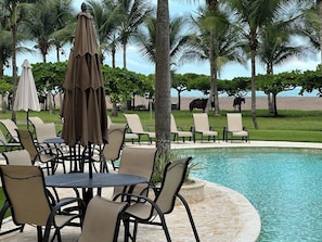 A poolside area with lounge chairs and umbrellas, surrounded by palm trees and a view of the sea. - Oceanfront Property Condo - 3 bedroom (Jaco)