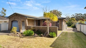 Une maison à un étage, construite en briques, avec un garage à l’avant, un petit jardin avec un arbre et des arbustes, et un ciel bleu dégagé.
