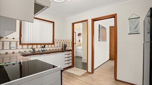 Une cuisine moderne avec des armoires blanches, un évier en acier inoxydable et un plan de travail en bois. On y trouve une fenêtre avec des stores, une porte donnant sur une autre pièce et un petit tapis sur le sol.