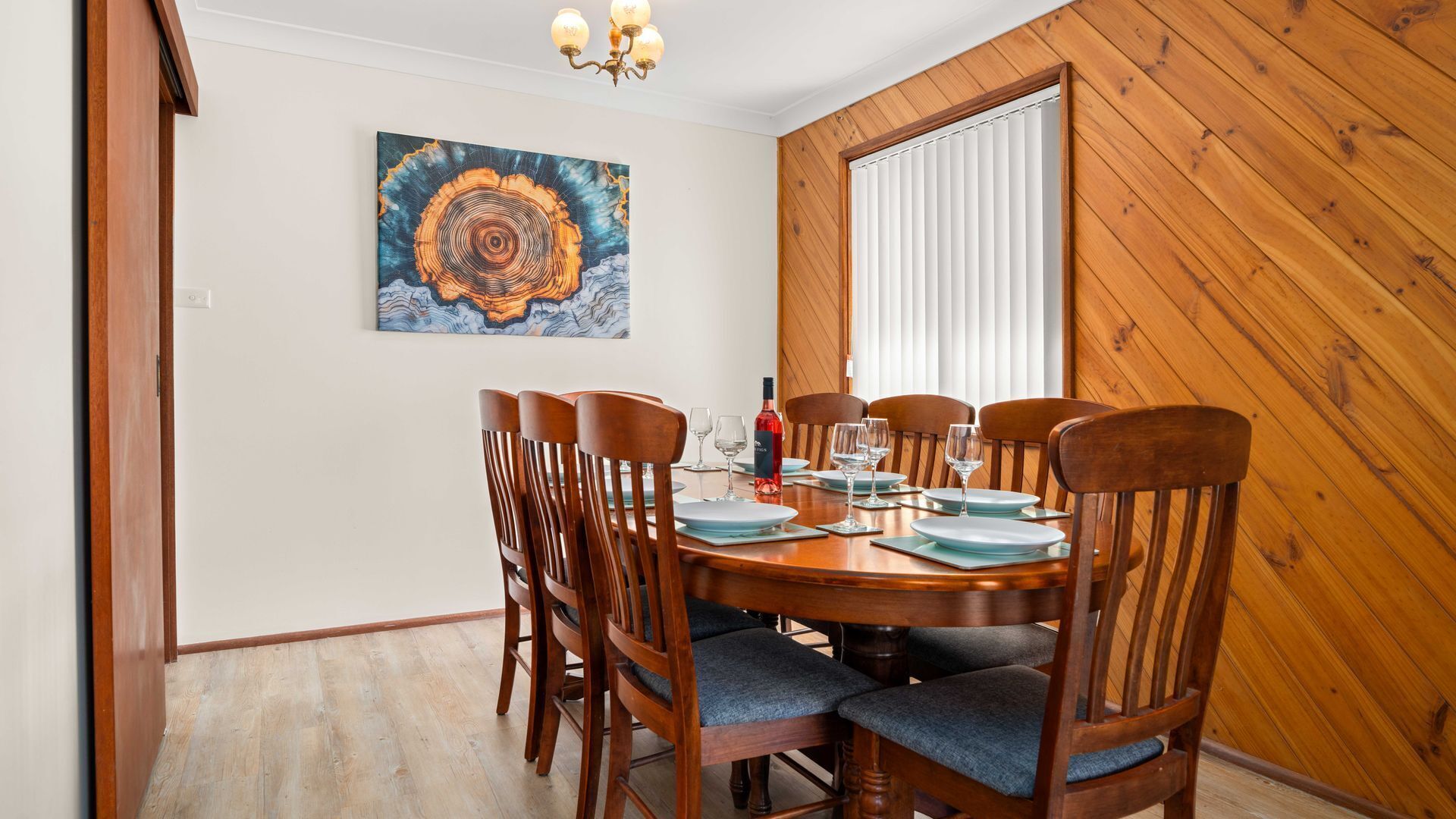 A dining room with a wooden table set for six, surrounded by six wooden chairs with blue cushions. A painting hangs on the wall above the table.