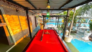 A pool table with a red felt surface, surrounded by a covered area with a view of a pool and outdoor plants. - Villa Na Wande Sattahip (Sattahip)