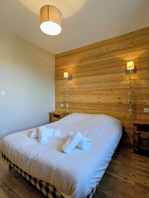 Una cama bien tendida con ropa de cama y toallas blancas, paneles de madera en las paredes y dos lámparas de pared.