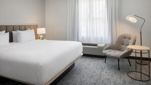 Habitación de hotel con una cama grande, una silla, una mesita, una lámpara y una ventana con cortinas traslúcidas.