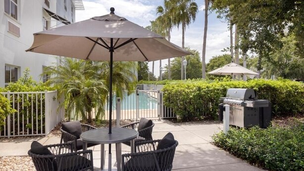 An outdoor patio with a round table and four chairs, a large umbrella, a grill, and a pool in the background. - Fully Equipped Kitchen in Modern Suite with Airport Shuttle and Swimming Pool (Tampa)