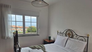 A neatly made bed with white linens and a green blanket, a bedside table with a green cup, and a window with a view of buildings and trees. - Charming apartment near the beach! Condominium with pool and playground (Amoreira)