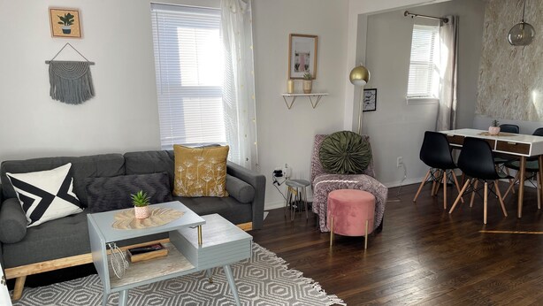 Una sala moderna con un sofá gris, una pequeña mesa de centro y un pouf rosa. Hay un comedor con una mesa blanca y sillas negras.