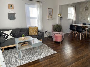 Una sala moderna con un sofá gris, una pequeña mesa de centro y un pouf rosa. Hay un comedor con una mesa blanca y sillas negras.