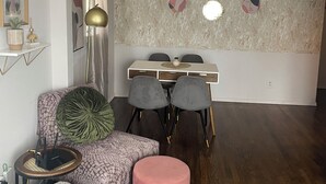 Una sala de estar moderna con suelo de madera, un sofá, un reposapiés rosa, una mesa de comedor con sillas, una lámpara de pie y una decoración de pared.