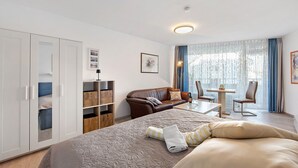 Habitación de hotel con cama, sofá, mesa de comedor, silla, armario y ventana grande con cortinas.