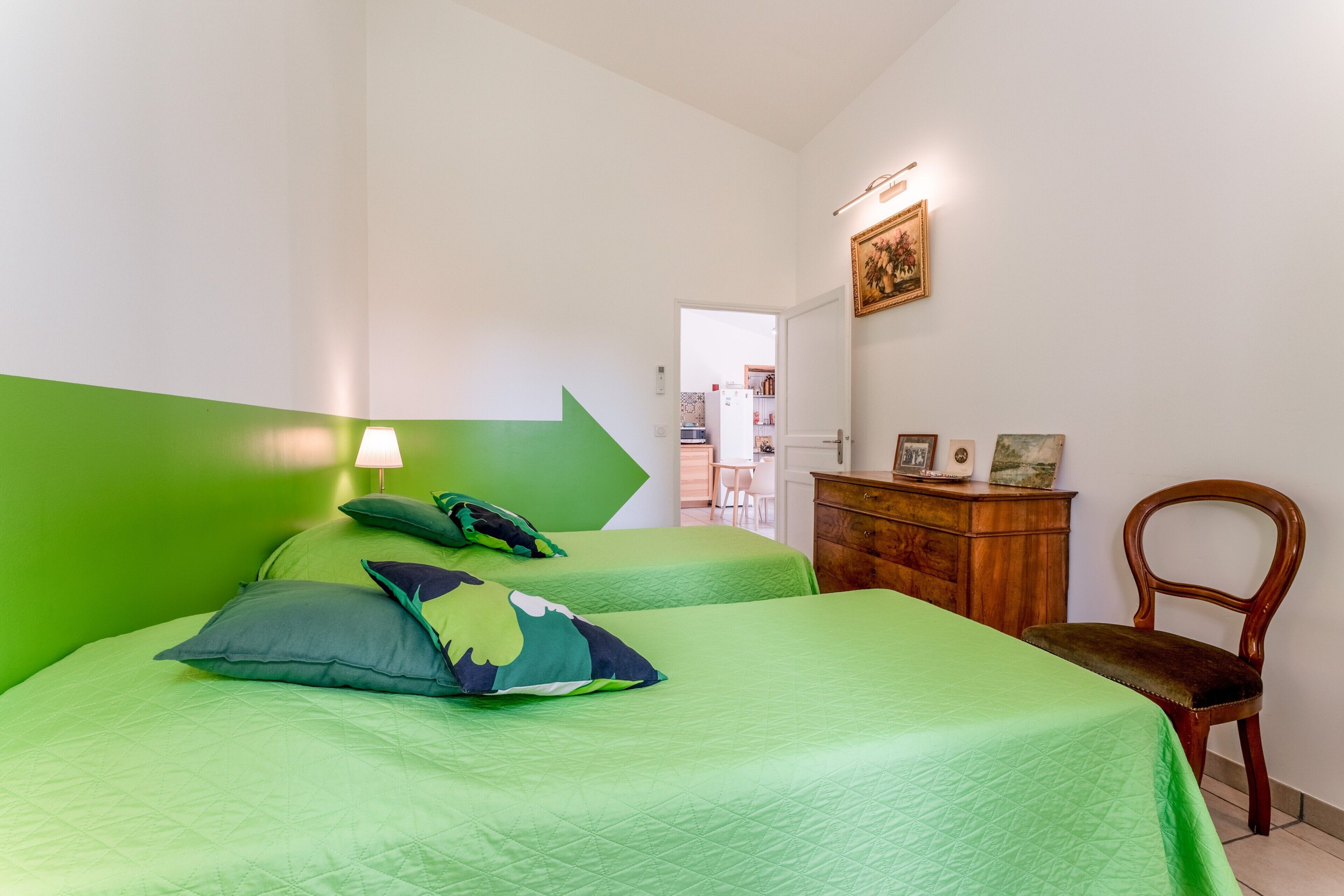 A room with two beds, a wooden dresser, a chair, and a door leading to another room.