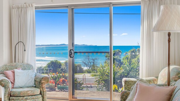 A room with a sliding glass door leading to a balcony with a view of the ocean and hills. There are two chairs with floral upholstery and a lamp on the right.