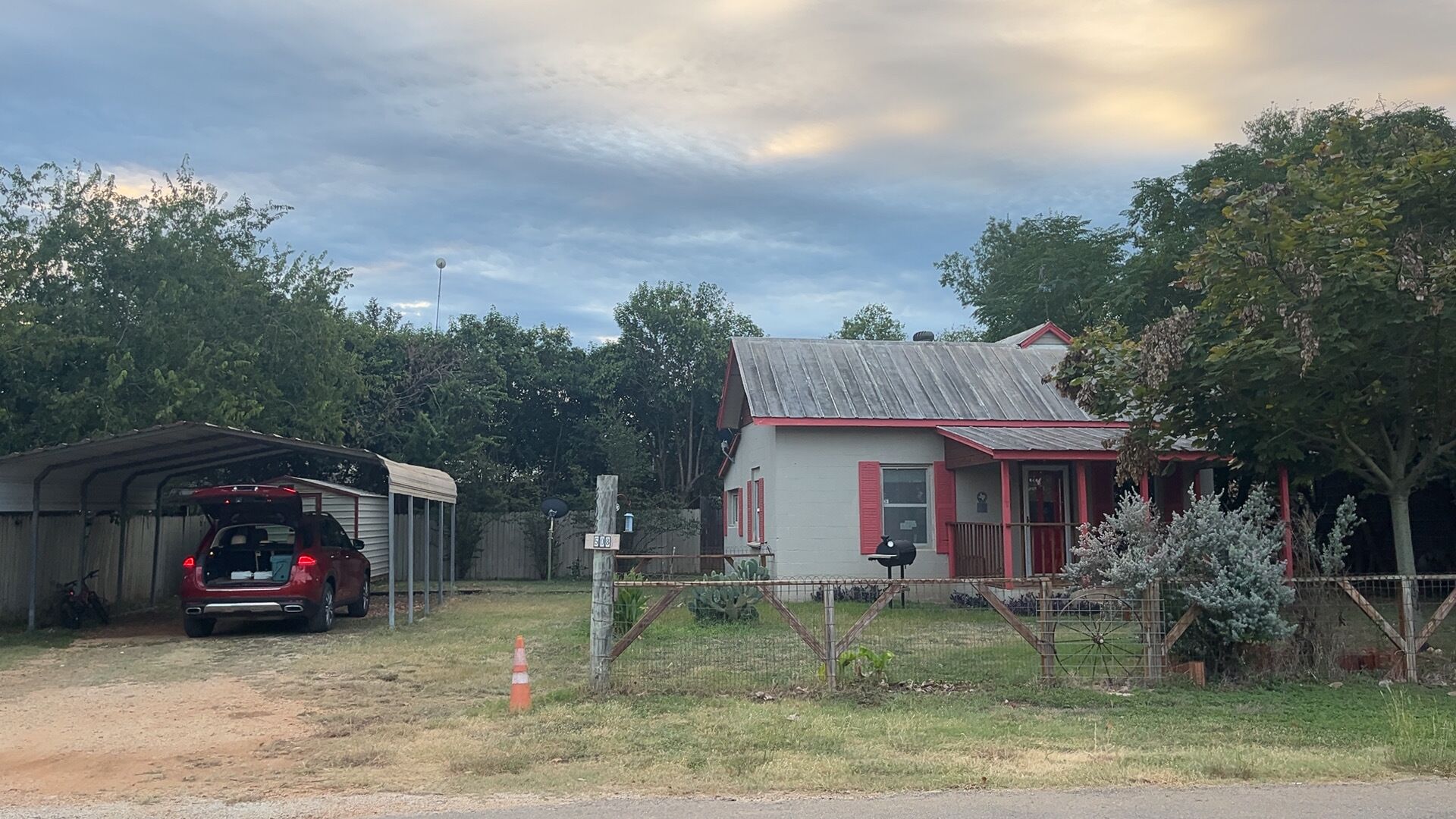 Paisagem rural com um carro vermelho estacionado numa garagem, uma casa pequena com telhado vermelho e um espaço cercado com uma árvore.
