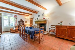 A dining area with a table set for four, wooden chairs, a fireplace, and a window with curtains. - Villa 'Le Mas' with Mountain View, Private Pool and Wi-Fi (Entrechaux)