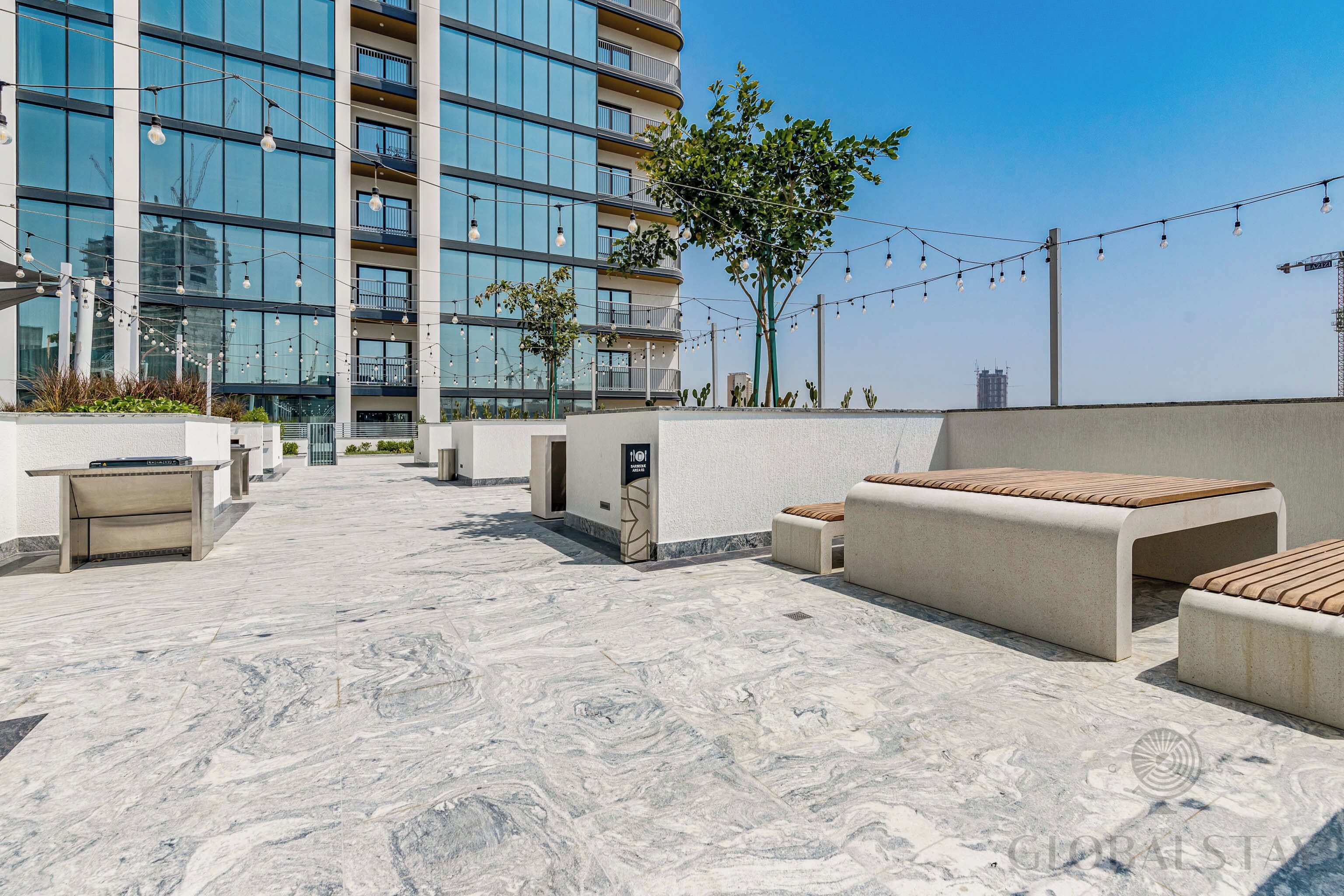 Una terraza en la azotea con mobiliario exterior, un edificio moderno al fondo y guirnaldas de luces.