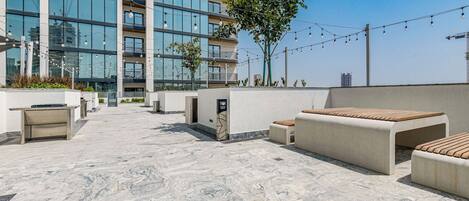 Una terraza en la azotea con mobiliario exterior, un edificio moderno al fondo y guirnaldas de luces.