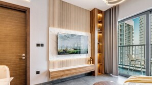 A modern living room with a large flat-screen TV mounted on a wooden panel wall, a built-in bench, and a balcony with a view of high-rise buildings. - GLOBALSTAY. The Crest Tower C Apartments (Dubai)
