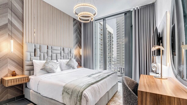 A modern bedroom with a large bed, a wooden nightstand, a desk, and a large window with curtains. - GLOBALSTAY. The Crest Tower C Apartments (Dubai)