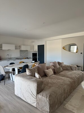 Ein modernes Wohnzimmer mit einem großen beigen Sofa, einem Esstisch mit Stühlen, einer Küchenzeile und einem großen Spiegel an der Wand.
