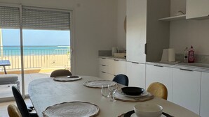 A dining area with a table set for four, a kitchen with white cabinets, and a view of the beach through large windows. - Holiday Home 'Face À La Mer 2' with Private Terrace and Air Conditioning (Port la nouvelle)