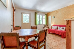 Un coin repas avec une table et des chaises en bois, un canapé recouvert d’une housse rouge et une fenêtre agrémentée de rideaux verts.