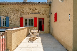 Une cour avec un mur en pierre, une table ronde entourĂ©e de chaises et des volets rouges aux fenĂȘtres.