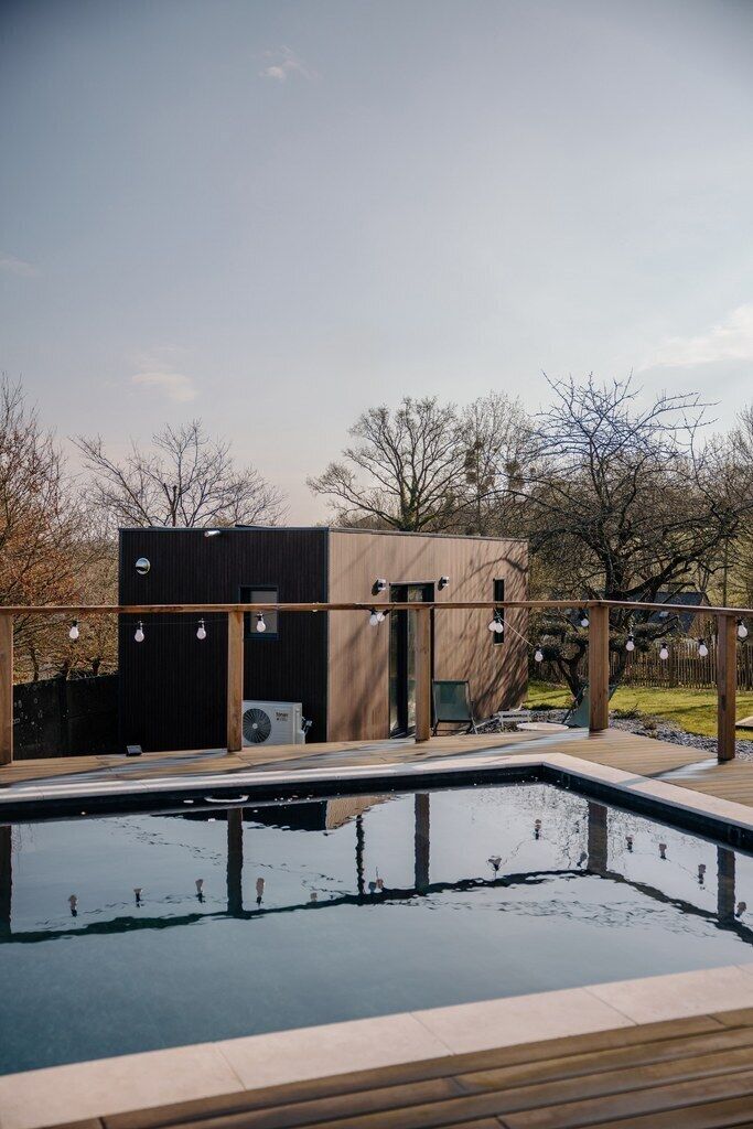 Une piscine avec une terrasse en bois, un bâtiment moderne à la façade noire et des arbres en arrière-plan.