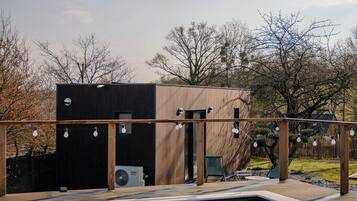 Une piscine avec une terrasse en bois, un bâtiment moderne à la façade noire et des arbres en arrière-plan.