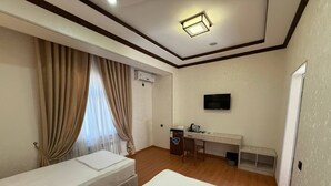 A hotel room with two beds, a desk with a chair, a TV mounted on the wall, and a window with curtains. - Pakhlavon Boutique Hotel (Khiva)