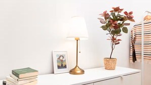 Ein weißer Nachttisch mit einer Messinglampe, einem gerahmten Bild, einem Bücherstapel und einer Topfpflanze.