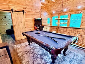 A pool table with cues, a television, and a hanging light fixture in a wooden room. - Luxury Cabin with Theatre and Hot Tub (Sevierville)