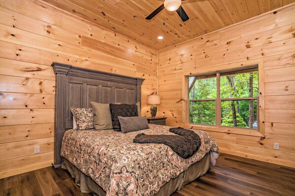 Un dormitorio con paneles de madera, una cama, almohadas, una mesita de noche, una lámpara y una ventana con vistas a árboles.