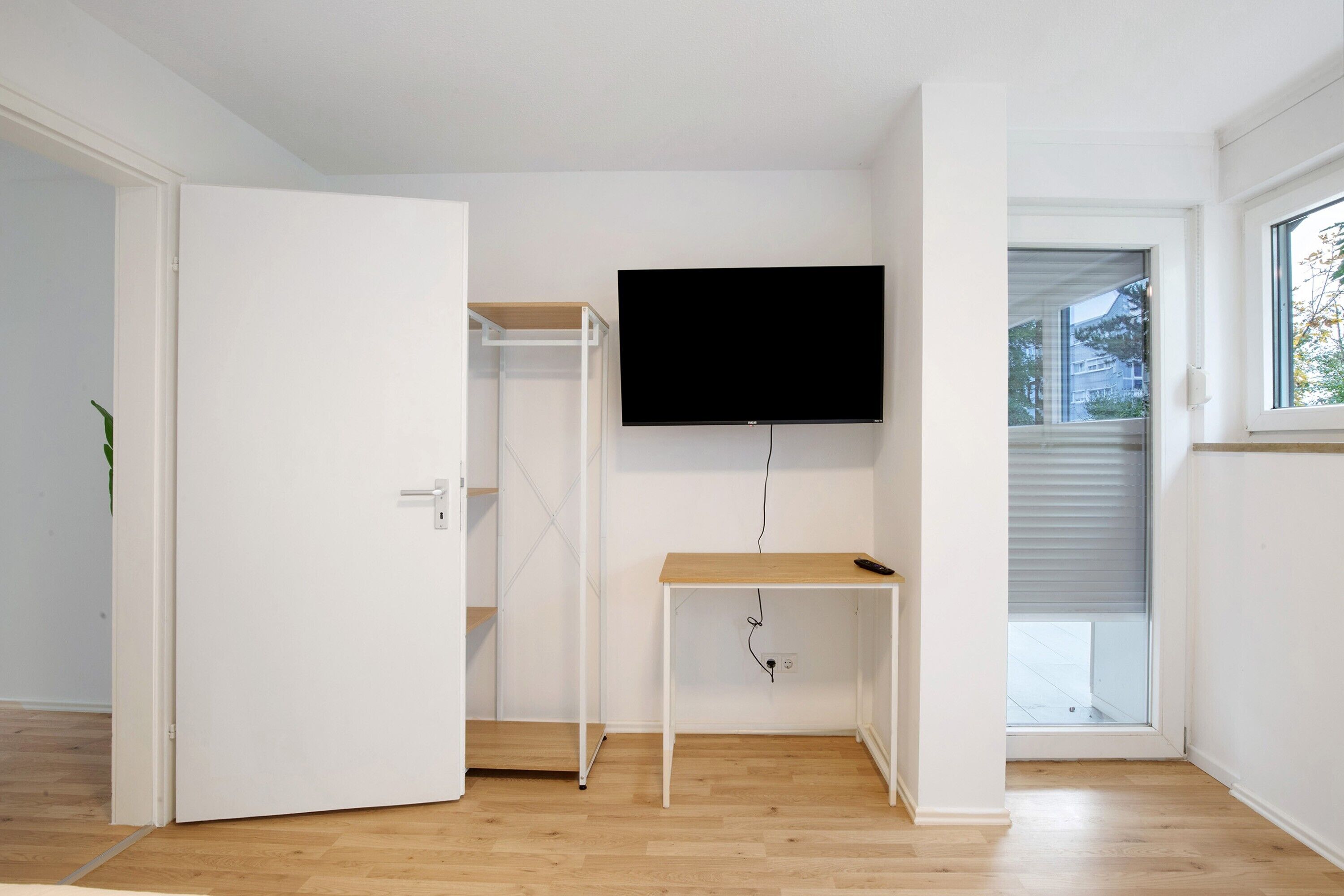 A room with a white door, a wooden floor, a white wall-mounted TV, and a window with blinds.