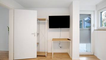 A room with a white door, a wooden floor, a white wall-mounted TV, and a window with blinds.